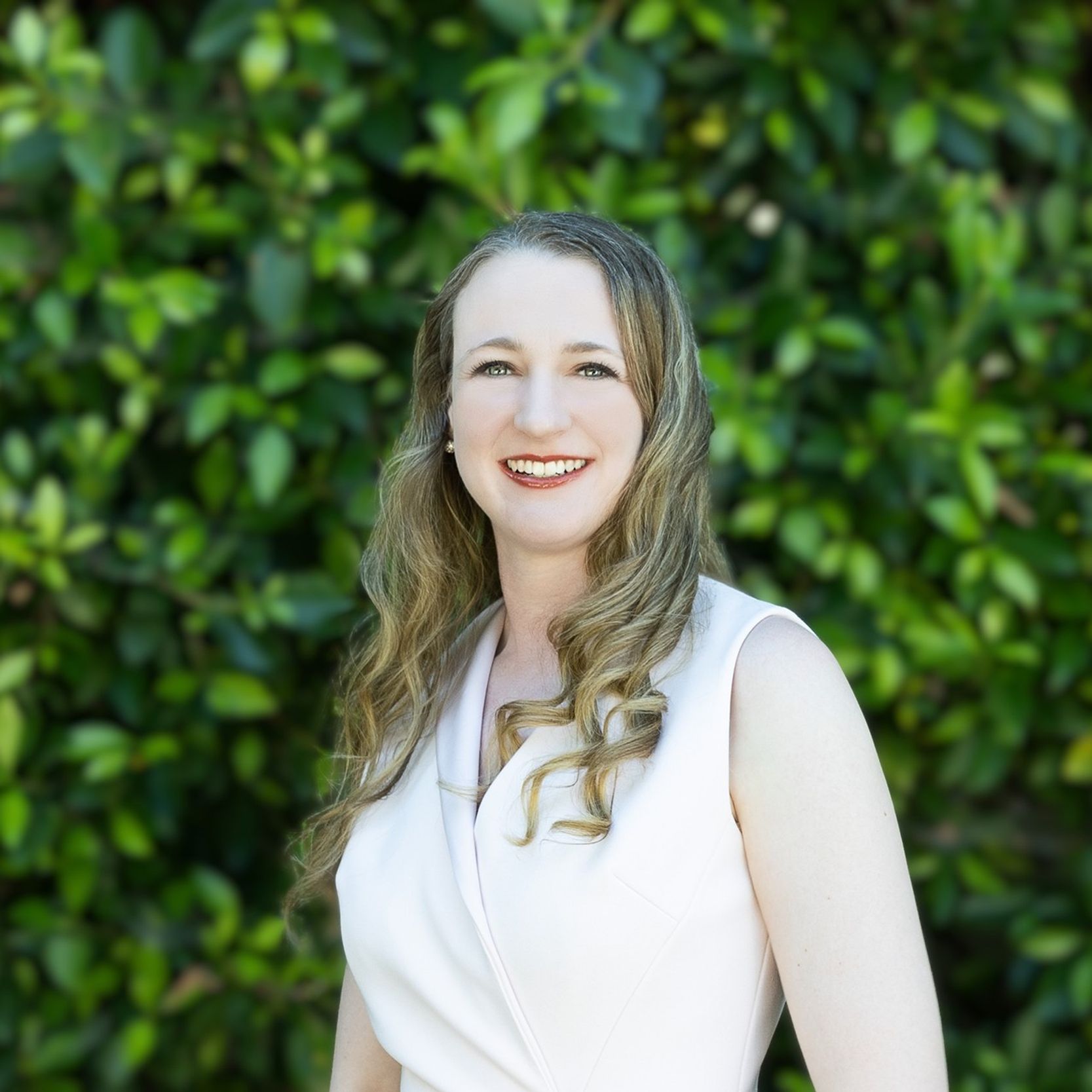 Portrait photo of a smiling woman wearing a white top and standing in front of greenery
