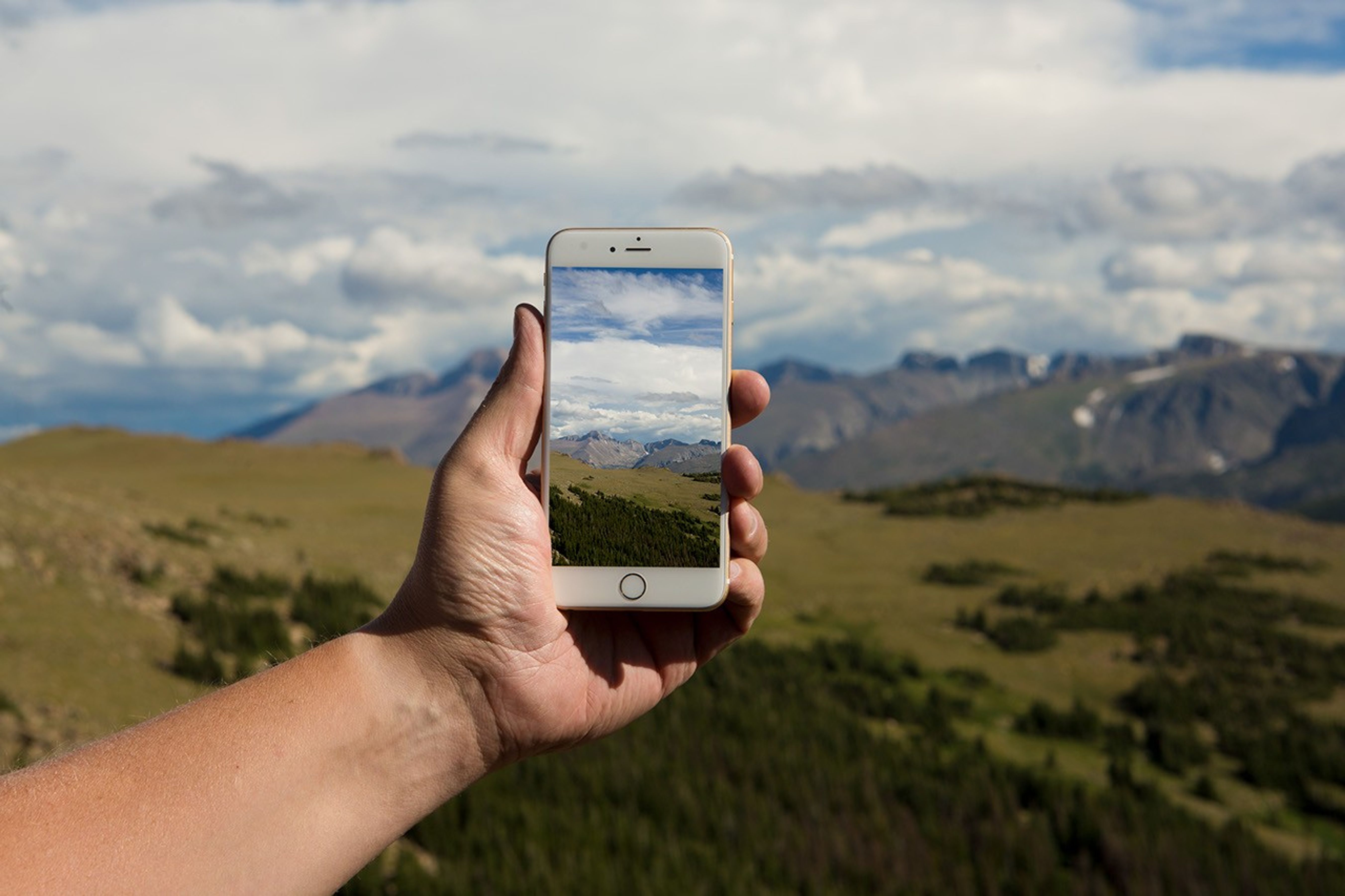 A hand holds a cell phone against a landscape of rolling hills and a cloudy sky.