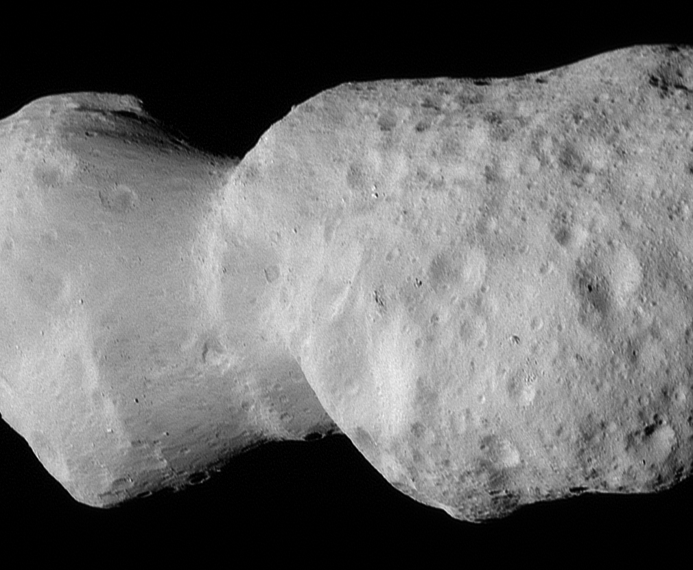 Asteroid Donaldjohanson, as seen by the Lucy spacecraft during its close flyby. The asteroid looks like two lobes, attached by a thinner cylinder, like a barbell with very large weights. One lobe is smaller than the other. Although they are both about the same circumference, the smaller lobe is less wide than the other. The asteroid is a smooth, light gray surface, with smooth-edged craters dimpling its surface. The larger lobe has many more craters.