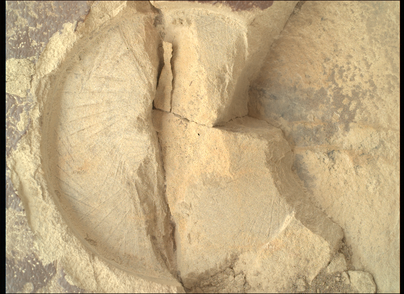 A color close-up photo from the Martian surface shows a circular impression on the flat, hard-looking stone surface, colored brownish-tan, which takes up almost all the image frame. The impression dimensions — its depth compared to its circumference — resembles that of a pie tin. The color of the terrain exposed in the impression is a pale warm tan, much lighter than the surrounding rock. The impression is fractured, in a crack straight down its middle. Another small crack points from the center toward the 4 o’clock position in the circle. To the right of that, the rest of the circle is cleaved off, and the entirety of about one-third of the right side of the circle is missing past a zig-zag line, running from about 1 o’clock to 5 o’clock, leaving an edge that descends below the surface, about twice the depth of the original impression.